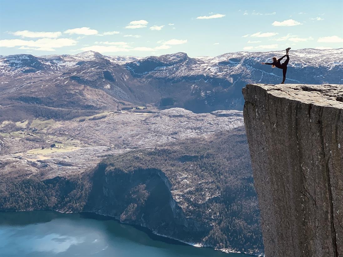 Das Bild zeigt eine Frau, die an einer steilen Klippe vor einer Berglandschaft mit einem See, eine Turnfigur macht.